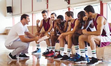 Male basketball coach explaining tactics to a team of players on a bench using a clipboard, illustrating the practical skills taught in the Sports Coaching Course.