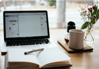Open laptop, blank notebook with pen, coffee cup, and flowers on a wooden desk near a window