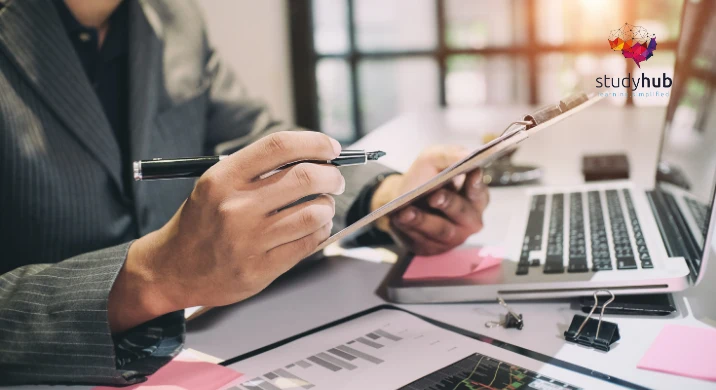 Professional bookkeeper in a suit reviewing financial graphs and data on a clipboard at a desk with a laptop, illustrating the Professional Bookkeeping Course.