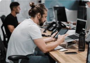 A man with a beard and hair bun sitting at a wooden desk in an office, holding a tablet while sitting in front of a laptop on a stand